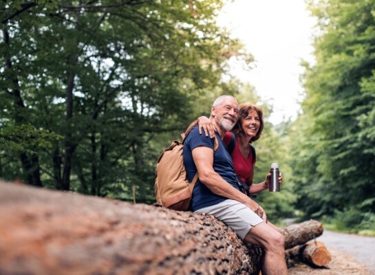 An older aged man and woman sitting arm and arm outdoors on a fallen tree.